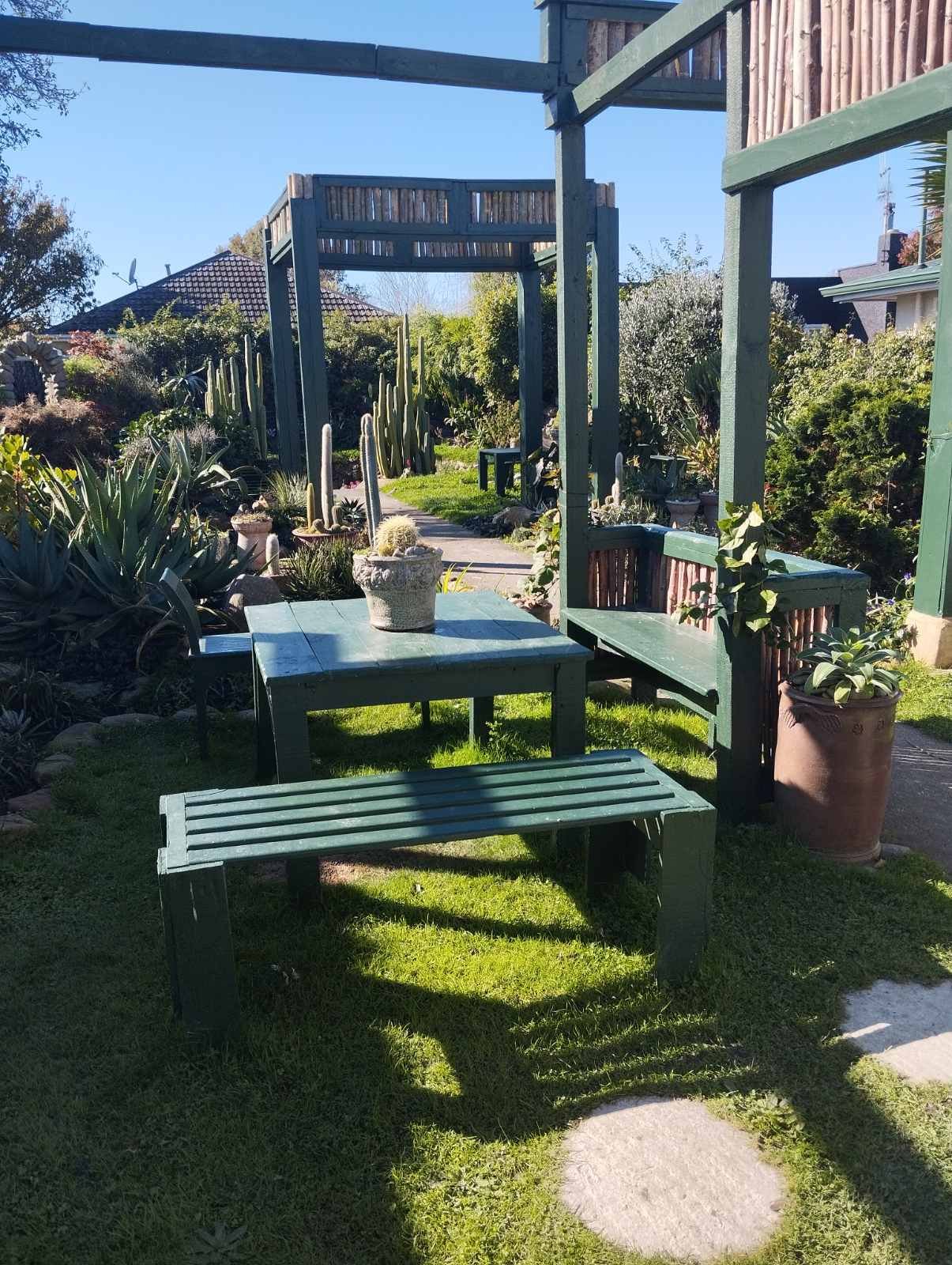 A sunny seating area in the Slo Mo Pots garden oasis, featuring a green wooden table and benches under a pergola, surrounded by cacti and succulents.