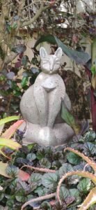 A serene, handcrafted ceramic cat sculpture with a textured grey glaze, sitting peacefully within a bed of purple and green garden plants.