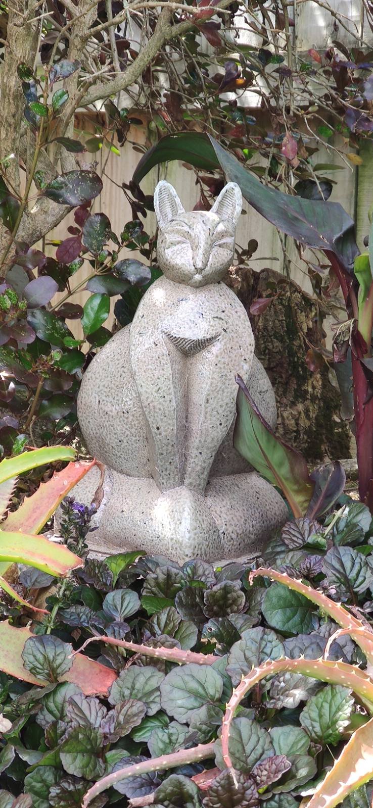 A serene, handcrafted ceramic cat sculpture with a textured grey glaze, sitting peacefully within a bed of purple and green garden plants.