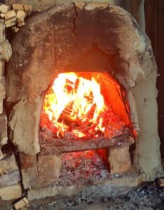 A close-up of the traditional wood-fired kiln at Slo Mo Pots, with bright orange flames and glowing embers roaring inside the rustic, earthen arch.