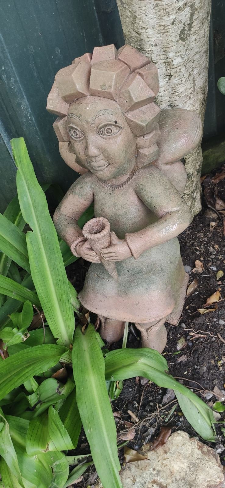 A whimsical, handcrafted clay sculpture of a figure with stylized hair, holding a small pot and standing by a tree in the garden.