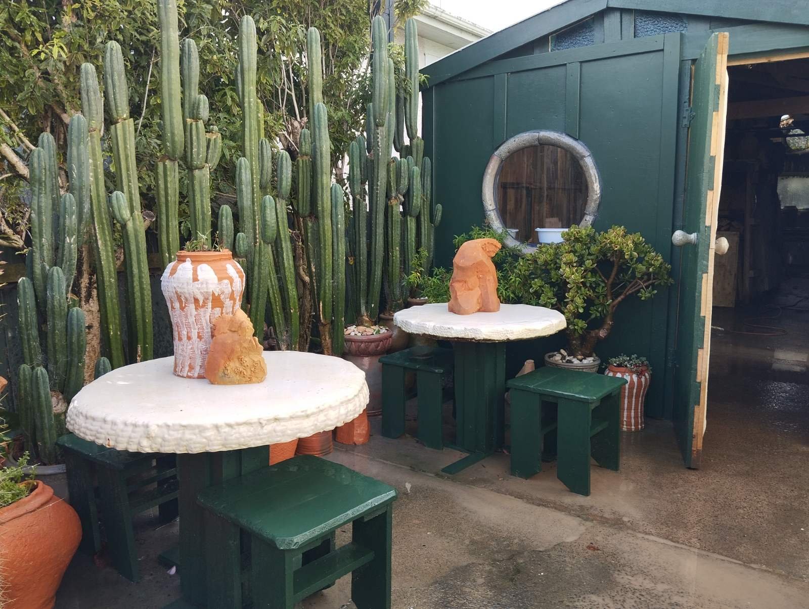 Outdoor seating area with clay sculptures on display, situated next to the green Slo Mo Pots studio building and a wall of tall San Pedro cacti.
