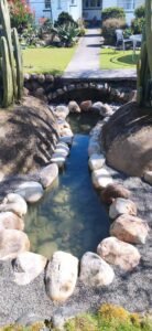 A tranquil, landscaped water feature in the garden, with clear water flowing through a channel lined with large, smooth river stones.
