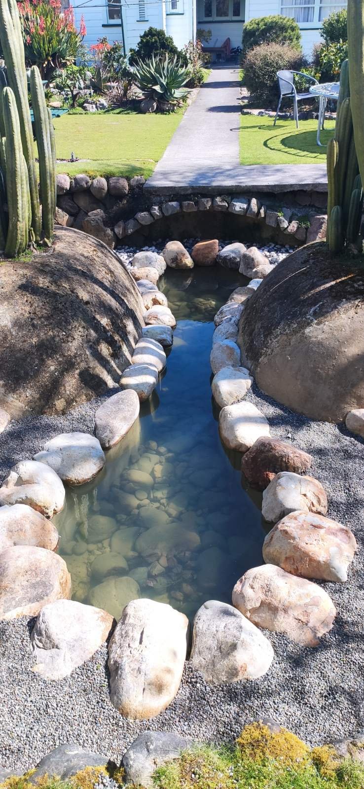 A tranquil, landscaped water feature in the garden, with clear water flowing through a channel lined with large, smooth river stones.