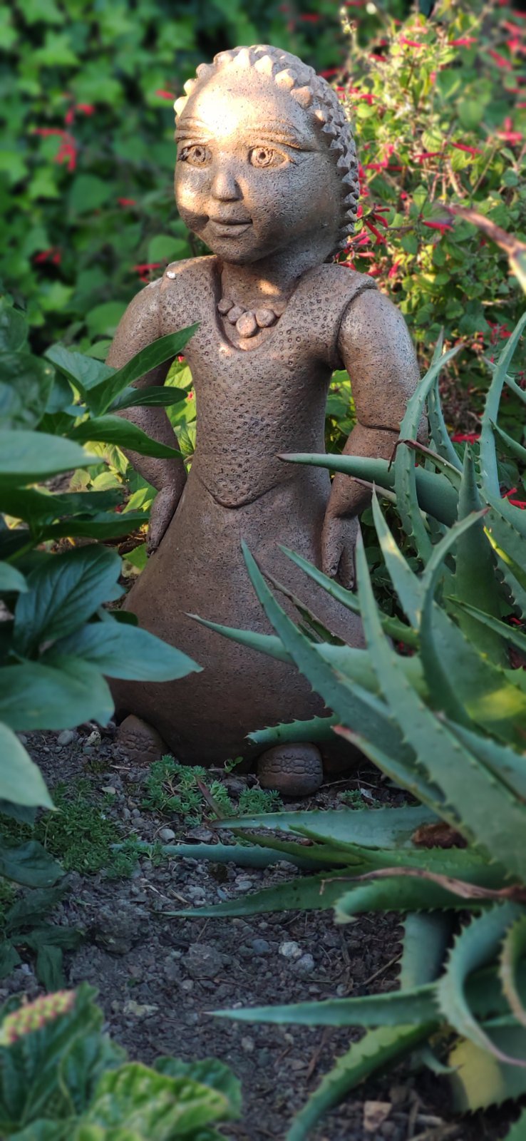 A handmade ceramic figure sculpture standing with hands on hips, artfully placed amongst aloe vera plants and other lush greenery.