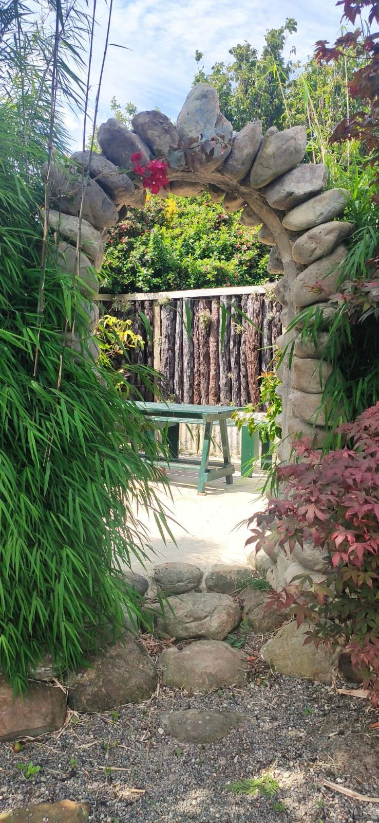 A charming stone archway in the garden, framed by lush bamboo and a Japanese maple, leading to a secluded seating area.