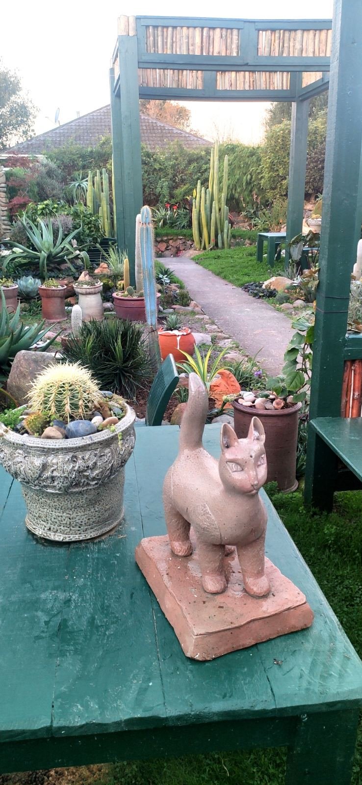 A handcrafted clay cat sculpture and a decorative cactus pot sitting on a green table in the Slo Mo Pots garden, with a view of the path.